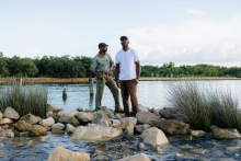 two men on rocks with water behind